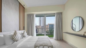 A modern hotel room with a large bed, a round mirror, and a balcony with a view of buildings. - Stylish 2BR w Study and City Views (Dubai Marina)