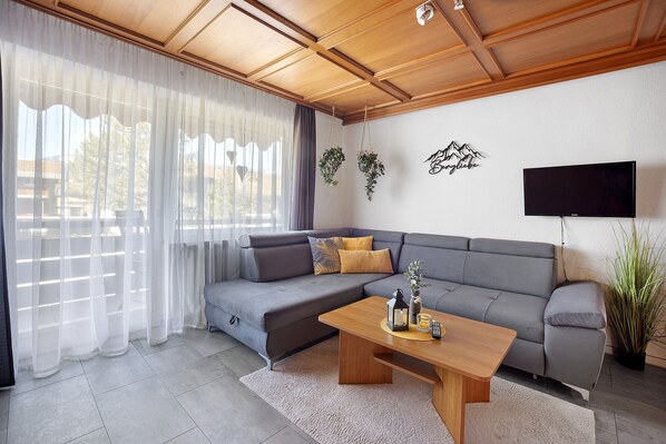 Un salon moderne avec un canapé d’angle gris, une table basse en bois, un téléviseur à écran plat et une plante en pot.