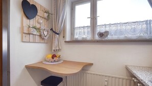Une cuisine avec une table en bois, un panier de fruits et une fenêtre ornée de rideaux en dentelle.