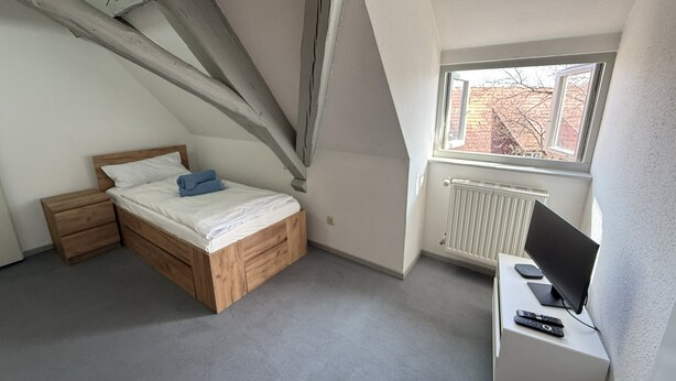 Une chambre à coucher avec un lit en bois, un bureau avec un ordinateur et une fenêtre donnant sur des immeubles.