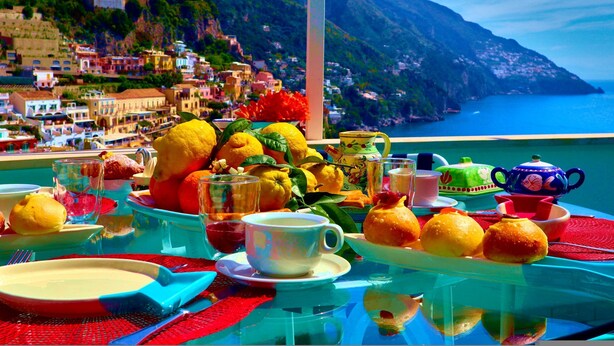 Uma mesa posta com frutas, um bule e xícaras, com vista para uma cidade costeira com edifícios e uma montanha.