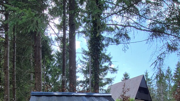 Ein Holzschild mit Schriftzug, ein Felsen und ein Haus in einer bewaldeten Gegend.