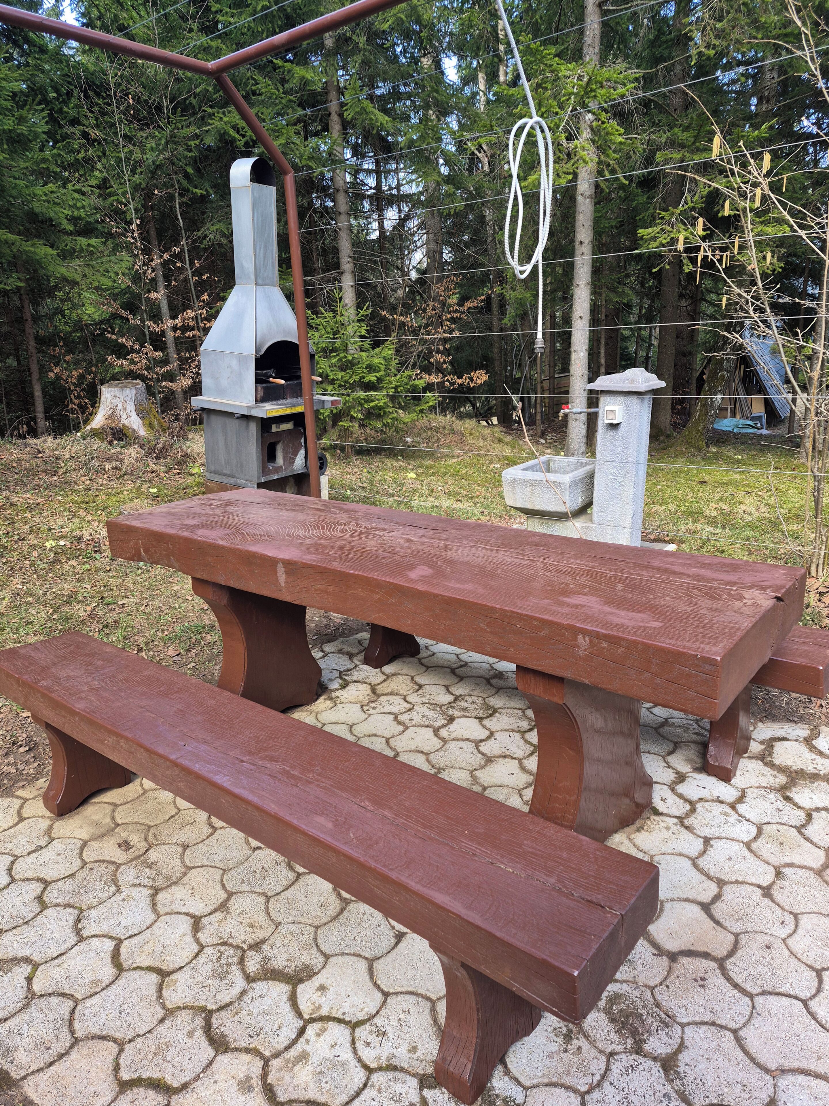 A picnic table with benches, a metal grill, and a stone structure in a wooded area.