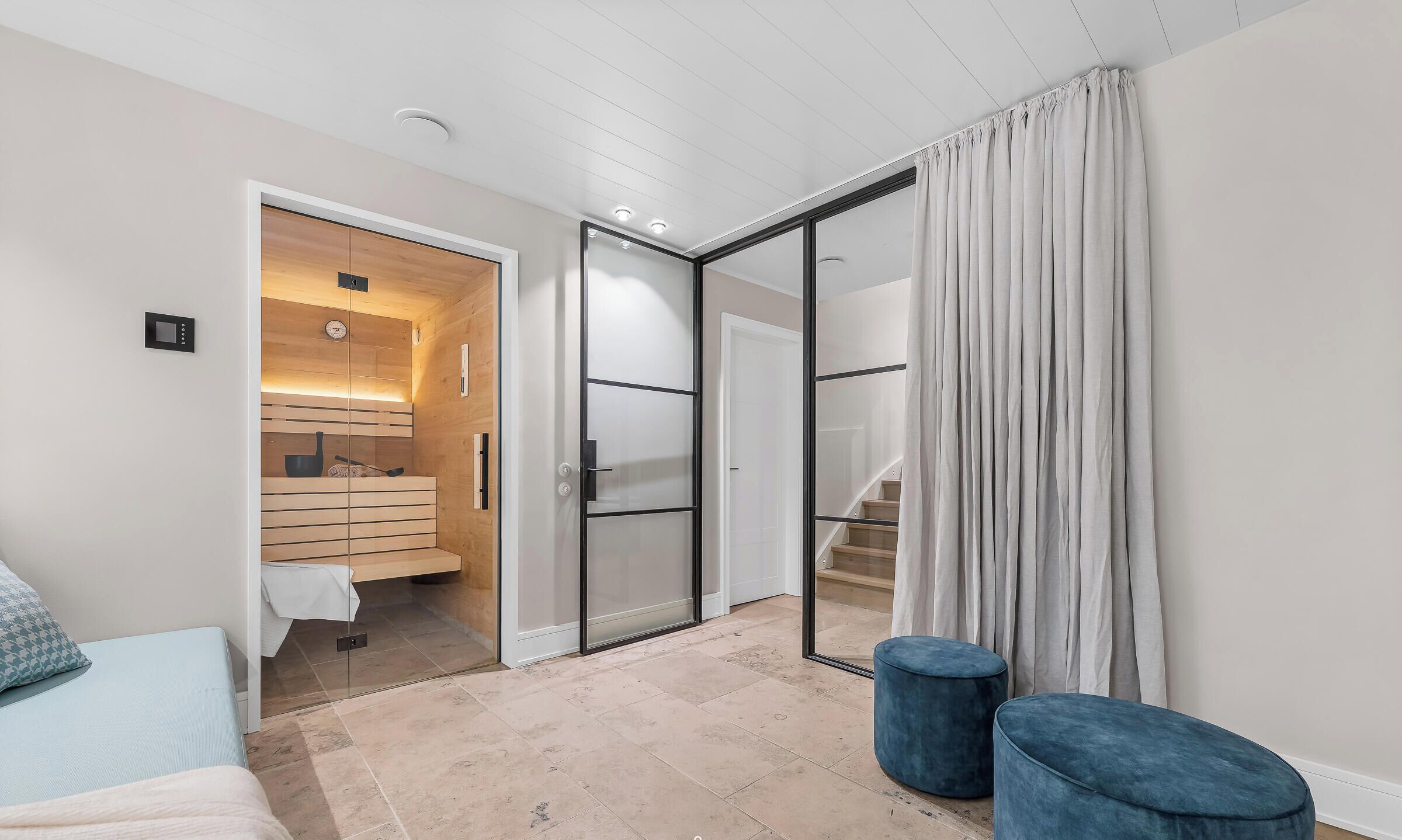 A modern room with a bed, a blue ottoman, a sliding glass door leading to a bathroom, and a staircase.