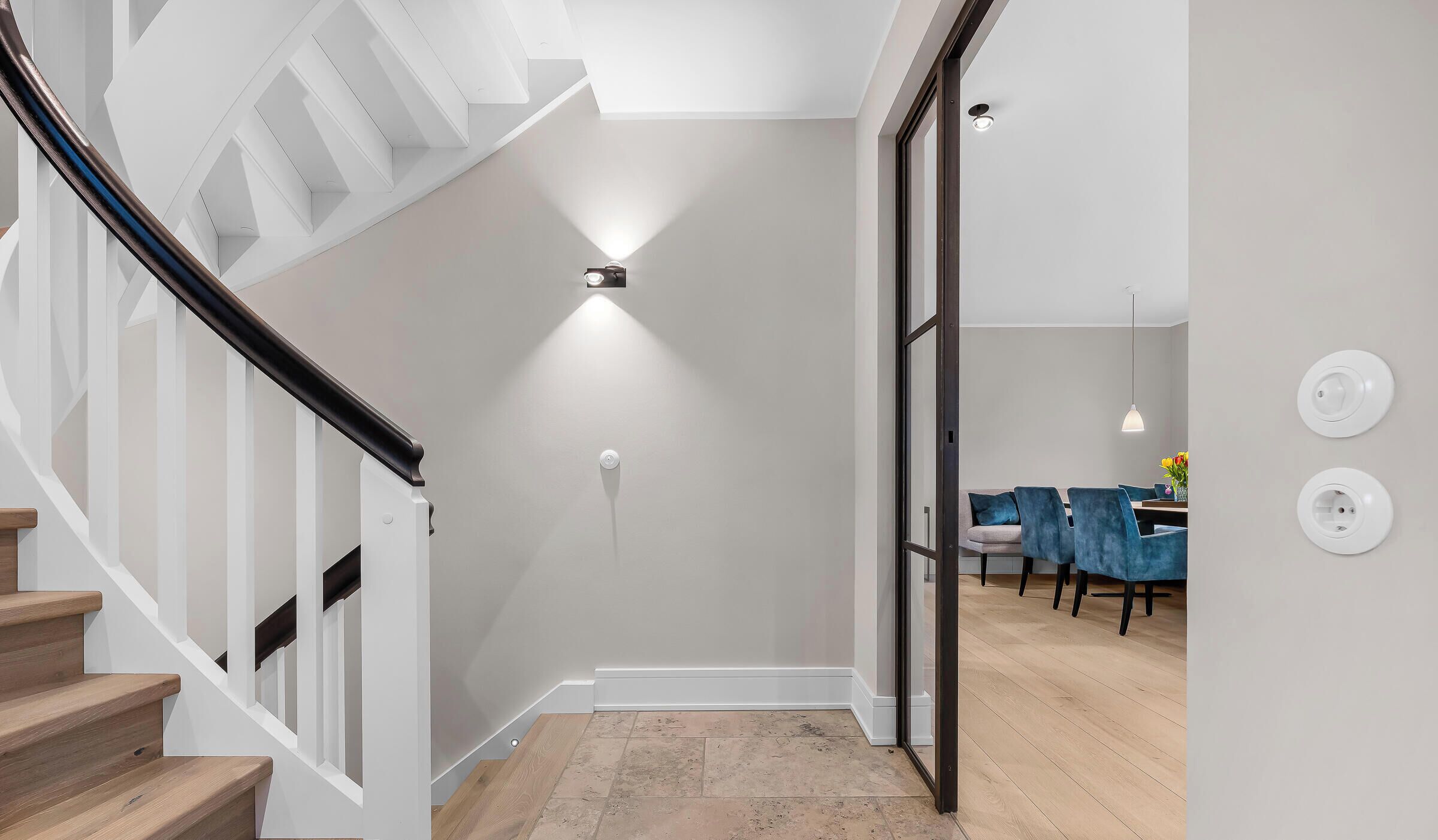 A modern interior with a staircase, a dining area with blue chairs, and a wall-mounted light fixture.