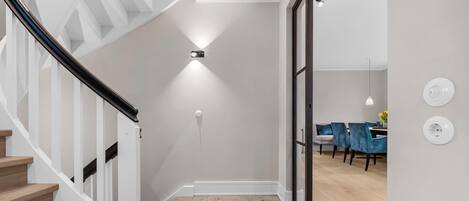 A modern interior with a staircase, a dining area with blue chairs, and a wall-mounted light fixture.