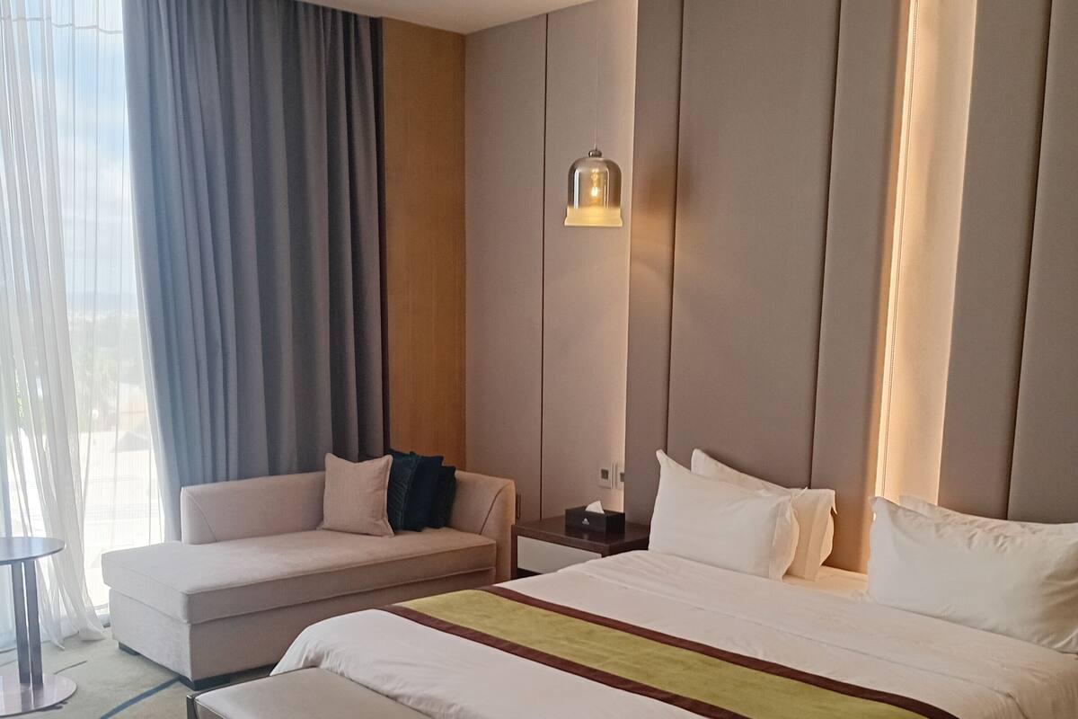 A hotel room with a large bed, a sofa, a bench, a bedside table, and a window with curtains.