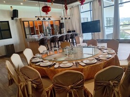 A conference room with a glass table, chairs with gold sashes, a bar area with glassware, and a large window offering a view.