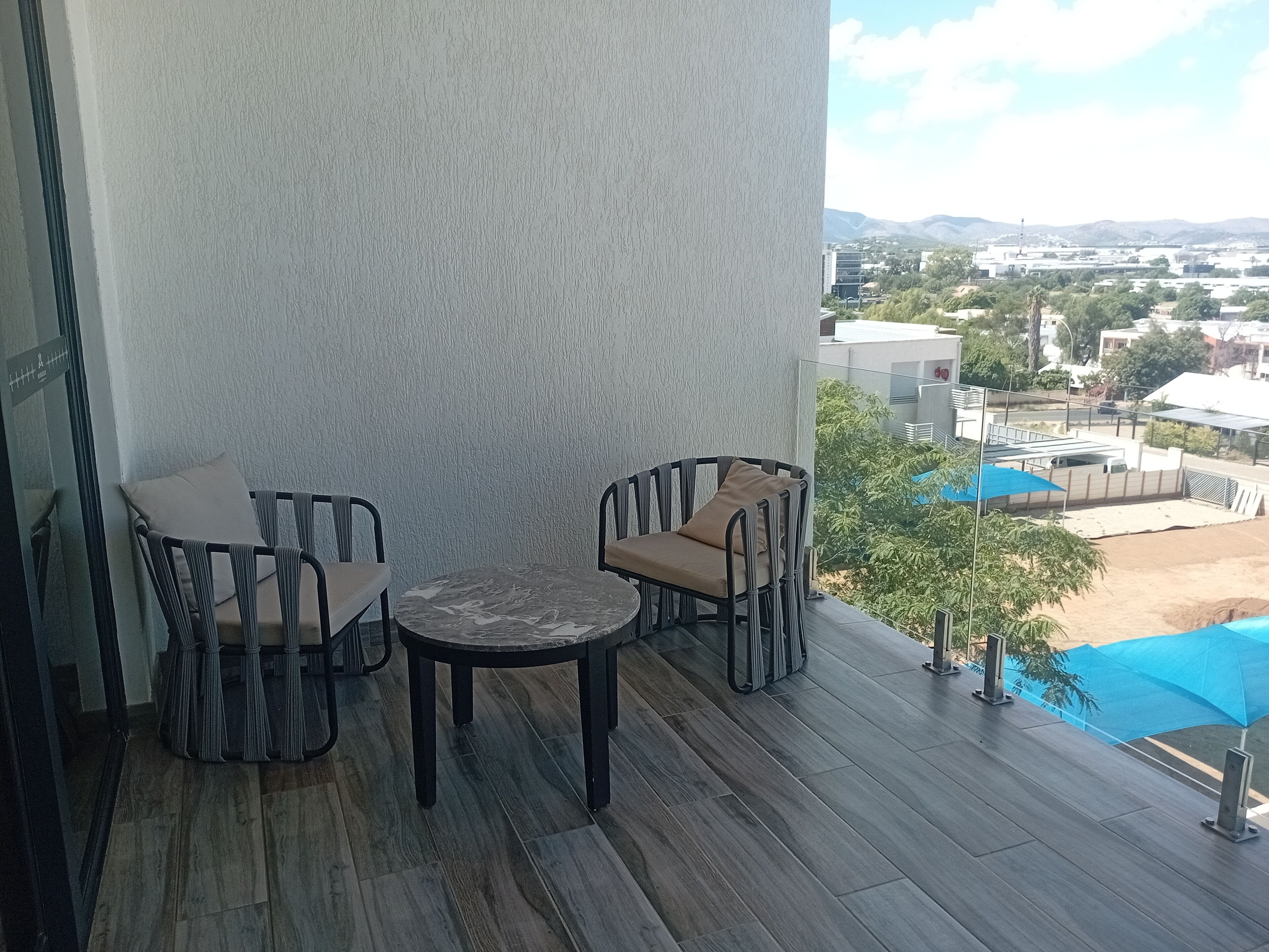 A balcony with two chairs and a table, a view of a pool and buildings in the background.