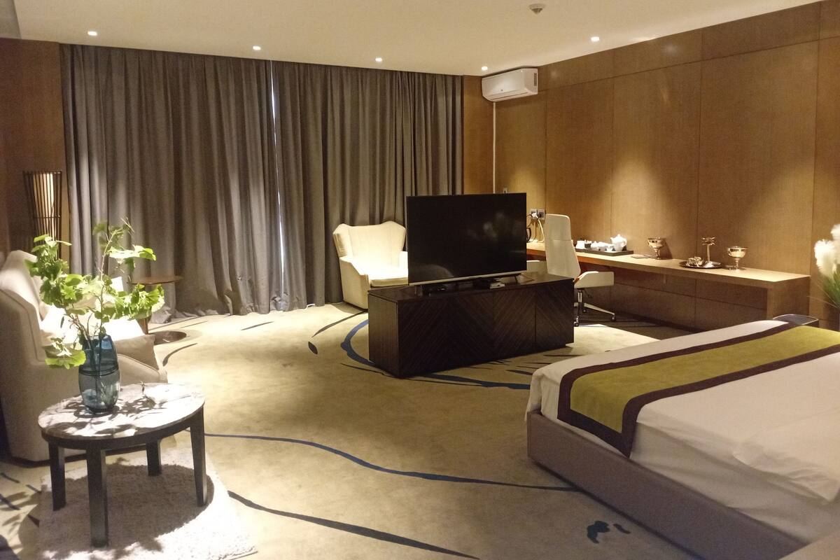 A hotel room with a bed, a desk, a television, a chair, a small table with a vase, and a curtain.