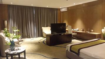 A hotel room with a bed, a desk, a television, a chair, a small table with a vase, and a curtain.