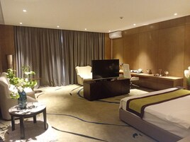 A hotel room with a bed, a desk, a television, a chair, a small table with a vase, and a curtain.