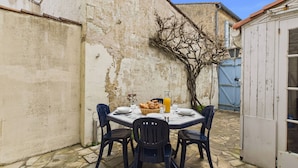 Un espace de restauration en plein air avec une table dressée pour deux personnes, entouré de carreaux de pierre, adossé à un mur patiné par le temps et agrémenté d’un arbre.
