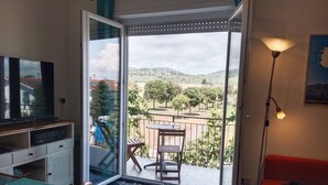 A room with a TV, a desk, a chair, a red sofa, a table, and a balcony with a view of trees and hills. - Casa Blu • Thermal Relax in the Center • Free Wi-Fi • 15 min. to the sea (Venturina Terme)