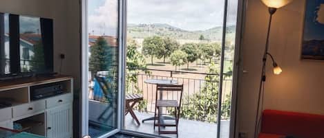 A room with a TV, a desk, a chair, a red sofa, a table, and a balcony with a view of trees and hills.