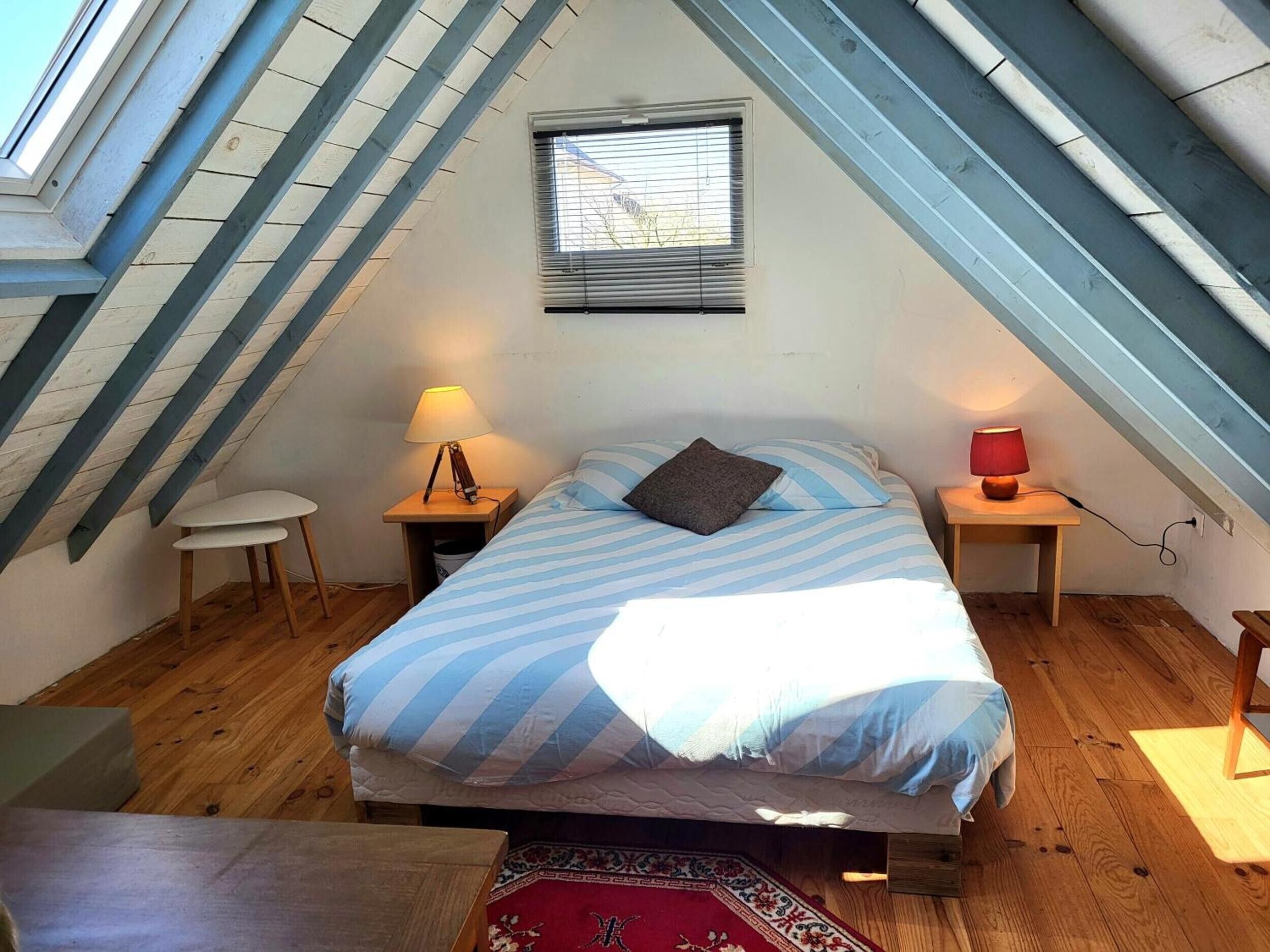 A bedroom with a sloped ceiling, a bed with striped bedding, two bedside lamps, a small table, and a window with blinds.