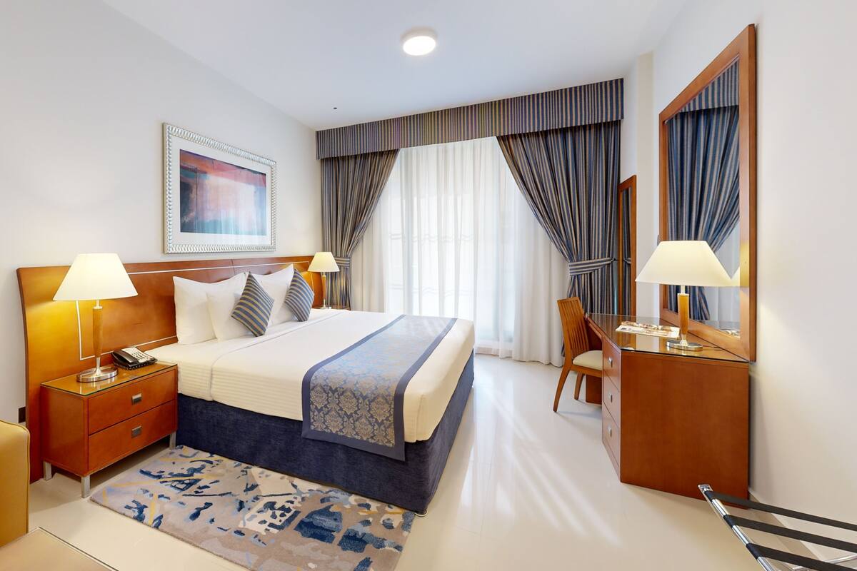 Habitación de hotel con cama, dos mesitas de noche con lámparas, un escritorio con silla, un espejo grande y un cuadro en la pared.