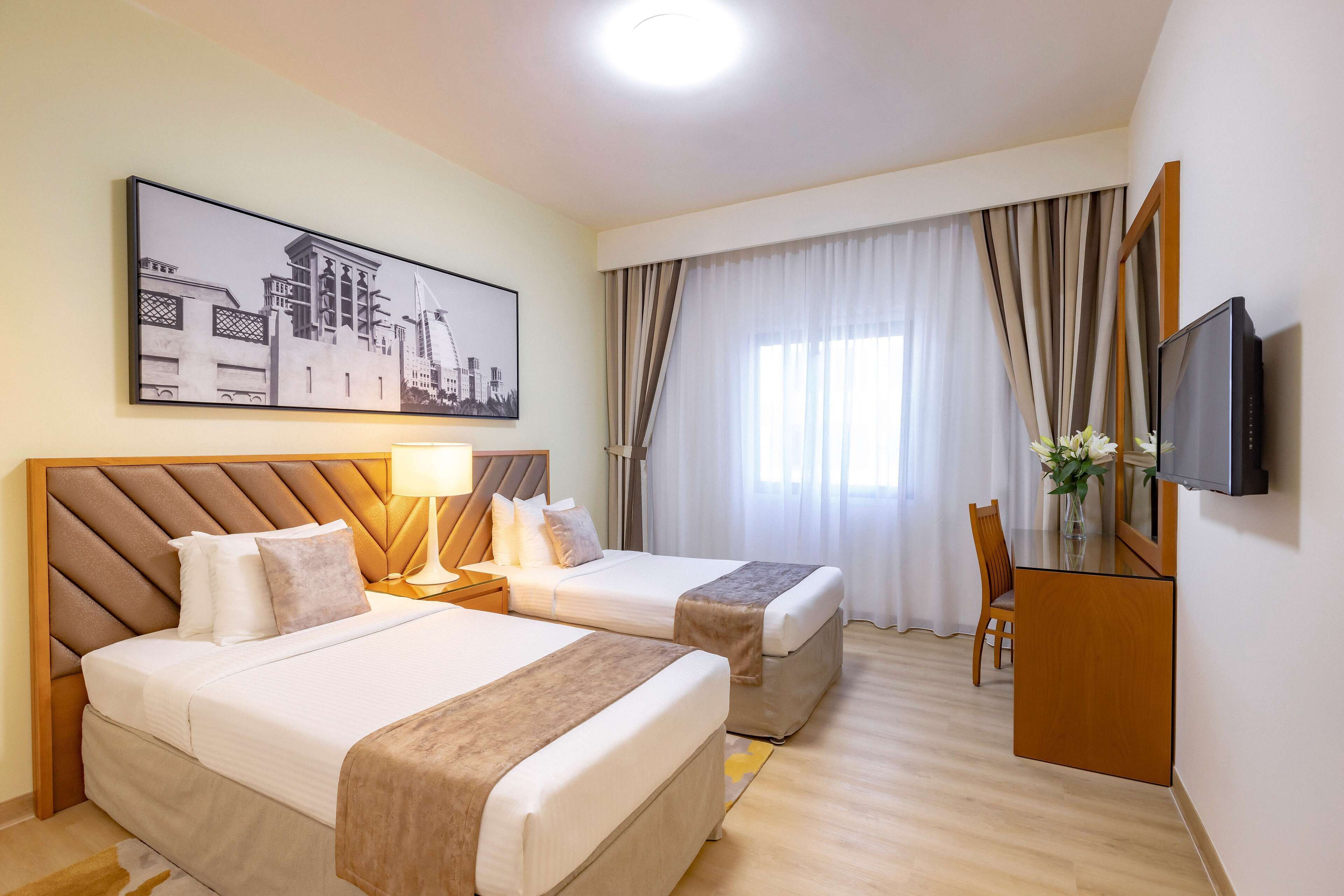 Habitación de hotel con dos camas, televisión, escritorio con silla y una ventana grande con cortinas.