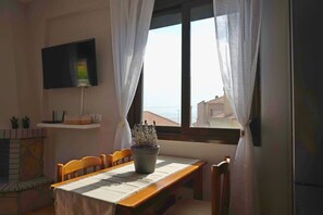 A dining area with a table, chairs, a potted plant, a flat-screen TV, and a window with curtains. - Into The Breeze by RentalsPro - Kriopigi Halkidiki (Kriopighi)