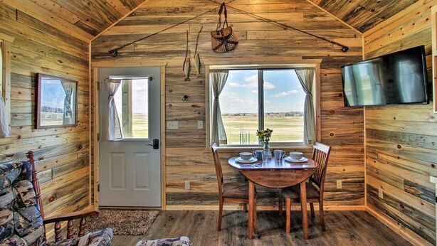A wooden cabin interior with a dining table, chairs, a TV, and fishing rods on the wall. - Stargazing Cabin Near Glacier w/ Loft & Fire Pit (Augusta)