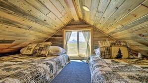 A cozy room with wooden walls and a sloped ceiling, featuring two beds with patterned bedding and a window offering a view of the outdoors. - Stargazing Cabin Near Glacier w/ Loft & Fire Pit (Augusta)