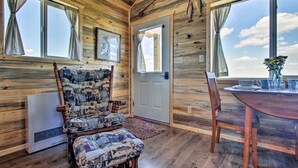 A wooden cabin interior with a patterned armchair, a dining table, and a view through a door. - Stargazing Cabin Near Glacier w/ Loft & Fire Pit (Augusta)