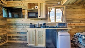 A compact kitchen with a microwave, toaster, and refrigerator, wooden walls, and a window with curtains. - Stargazing Cabin Near Glacier w/ Loft & Fire Pit (Augusta)