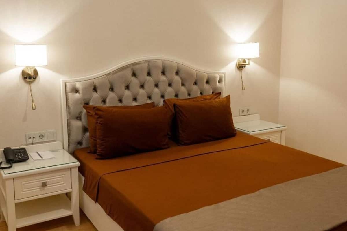 Uma cama com cabeceira estofada, duas luminárias de cabeceira, um telefone ao lado da cama e um espalha cama marrom.