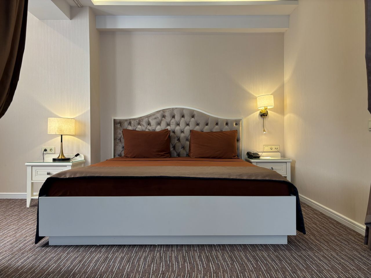 Una habitación de hotel con una cama grande, dos mesitas de noche con lámparas y piso alfombrado.