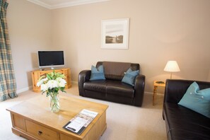 A living room with a flat-screen TV, a leather sofa with cushions, a coffee table with a magazine, and a lamp. - Holstein Cottage - The Dairy at Daviot (Daviot)
