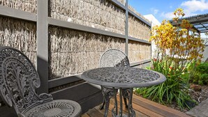 Une terrasse avec une table ronde et deux chaises décoratives, un mur orné et un jardin parsemé de fleurs jaunes.