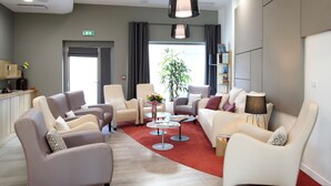 A modern hotel lobby with leather sofas, a coffee table, a bookshelf, and a large window with curtains. - Zenao Appart'hôtels Massy (Massy)