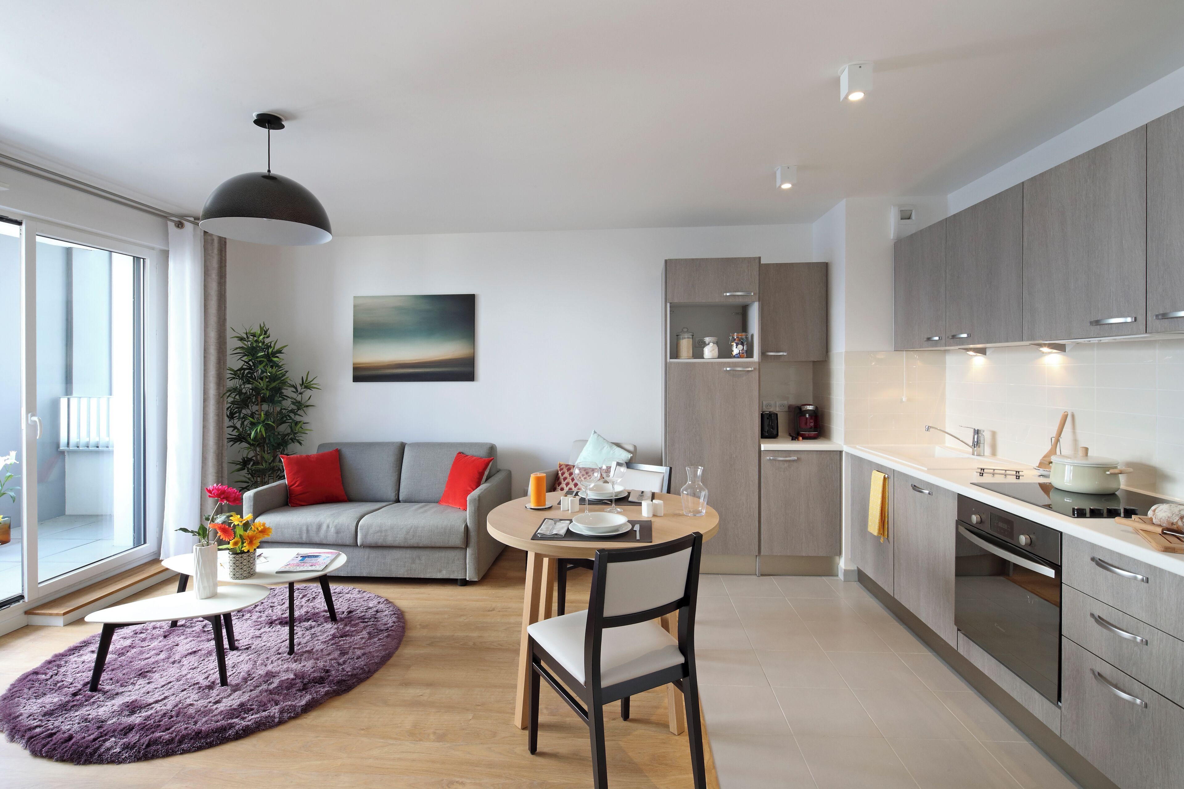 A modern kitchen with grey cabinets, a dining area with a round table and chairs, a grey sofa with red cushions, and a large window with a view.