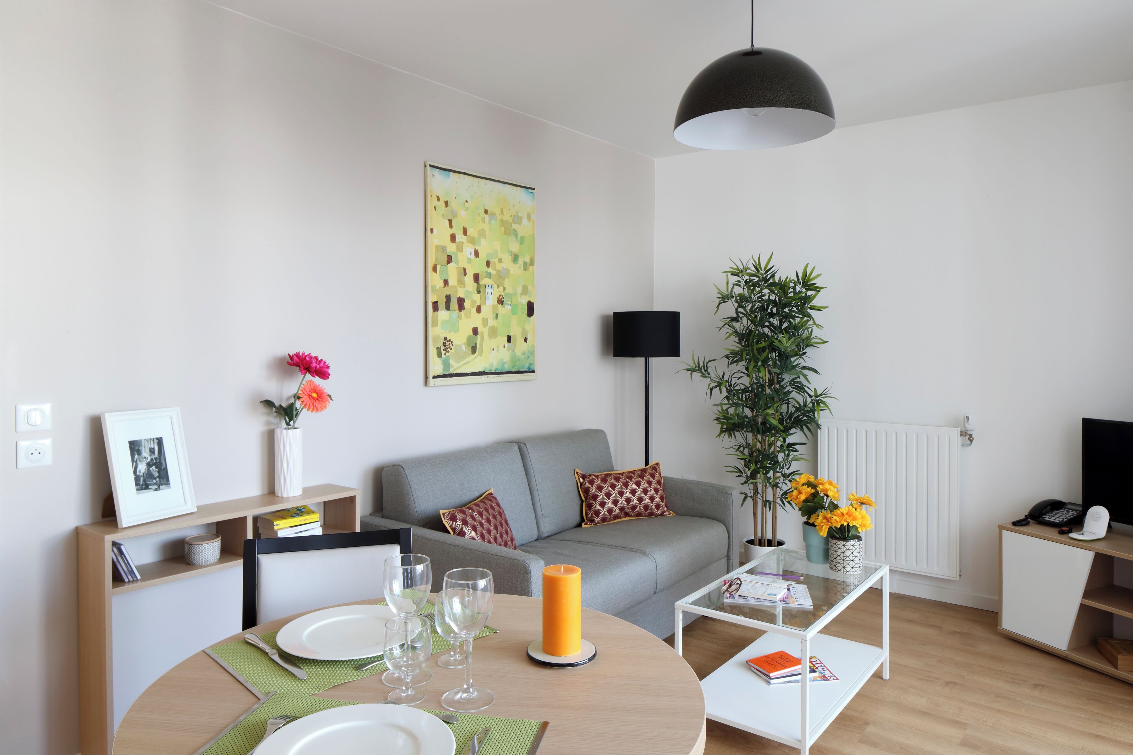 A modern living room with a grey sofa, a round dining table with plates and glasses, a black pendant light, a potted plant, and a framed picture on a shelf.