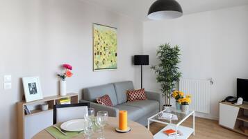 A modern living room with a grey sofa, a round dining table with plates and glasses, a black pendant light, a potted plant, and a framed picture on a shelf.