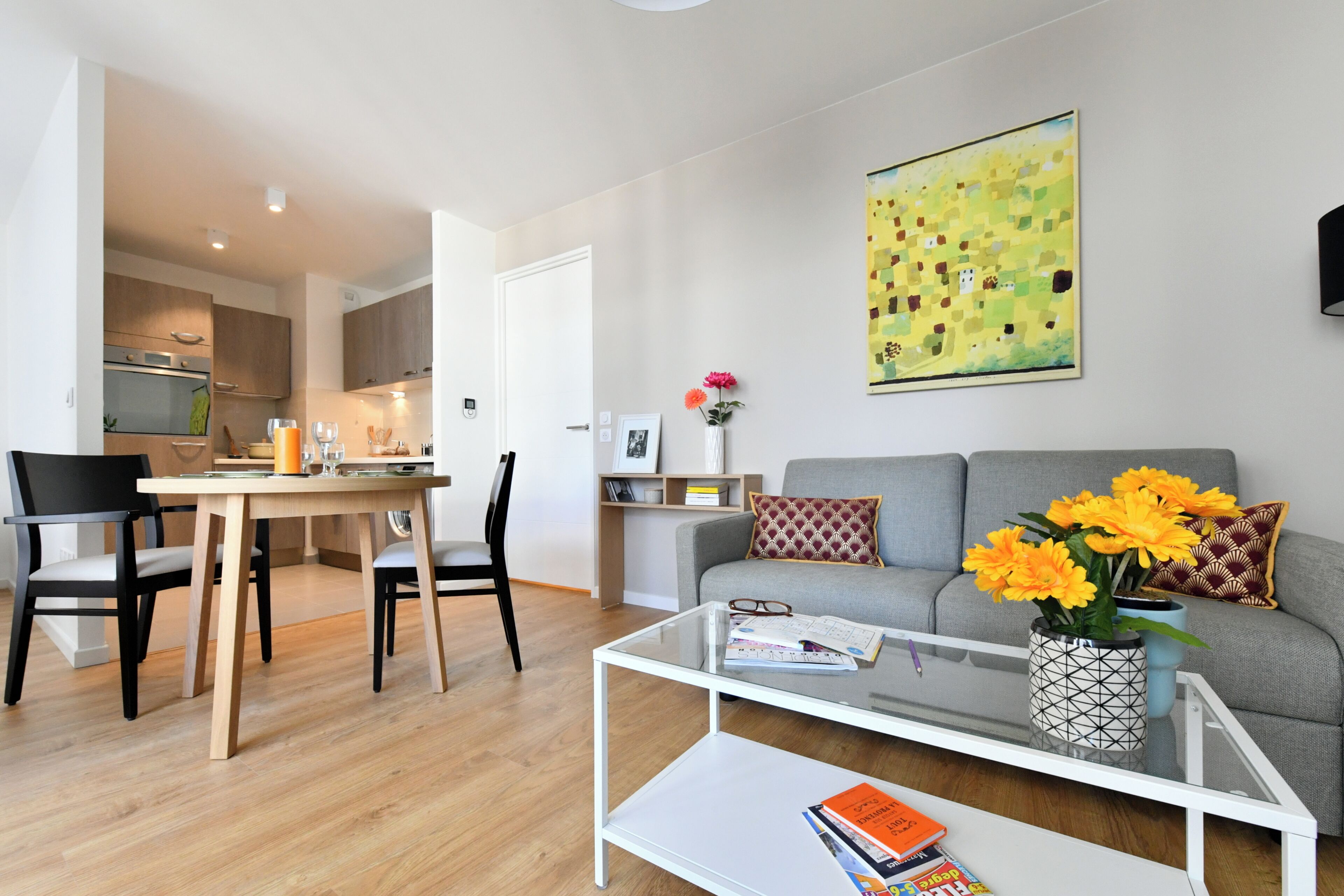 Un salon moderne avec une table basse en verre, un canapé gris orné de motifs floraux orange, un coin repas avec une table et des chaises en bois, et une cuisine en arrière-plan.