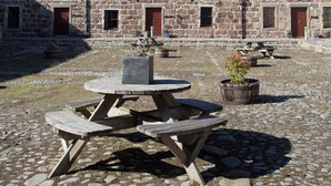 Ein Steingebäude mit zahlreichen Fenstern und Türen, ein Innenhof mit Picknicktisch und Blumentöpfen, und ein strahlend blauer Himmel.