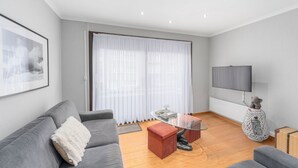 Een moderne woonkamer met een grijze bank, een glazen salontafel, een rode poef en een aan de muur bevestigde televisie.
