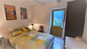 A bedroom with a bed, a nightstand, a lamp, a chair, a window with a view, and two posters on the wall. - In a quiet, green setting, pretty Provencal country house (Annot)