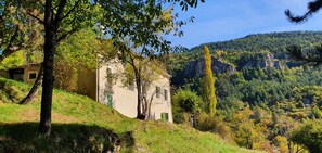 A rustic building with a sloped roof, surrounded by lush greenery and a mountainous backdrop. - In a quiet, green setting, pretty Provencal country house (Annot)