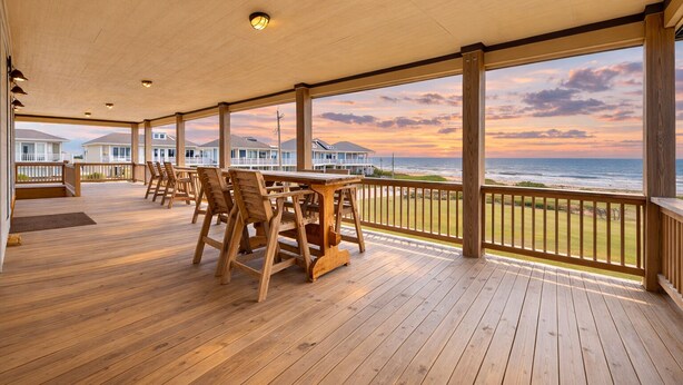 Eine Holzterrasse mit Tisch und Stühlen, die auf einen Strand und den Ozean bei Sonnenuntergang blickt.