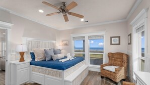 Ein Schlafzimmer mit Bett, Sessel, Fenster mit Blick, Deckenventilator und einem Wandbild.