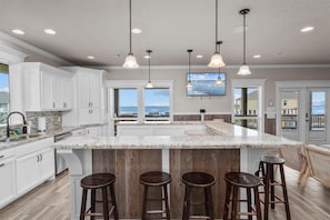 A kitchen with white cabinets, a granite countertop, wooden stools, and pendant lighting. - Beachfront | 6BR | Sleeps 33 | Hot Tub | Pickleball (Crystal Beach)