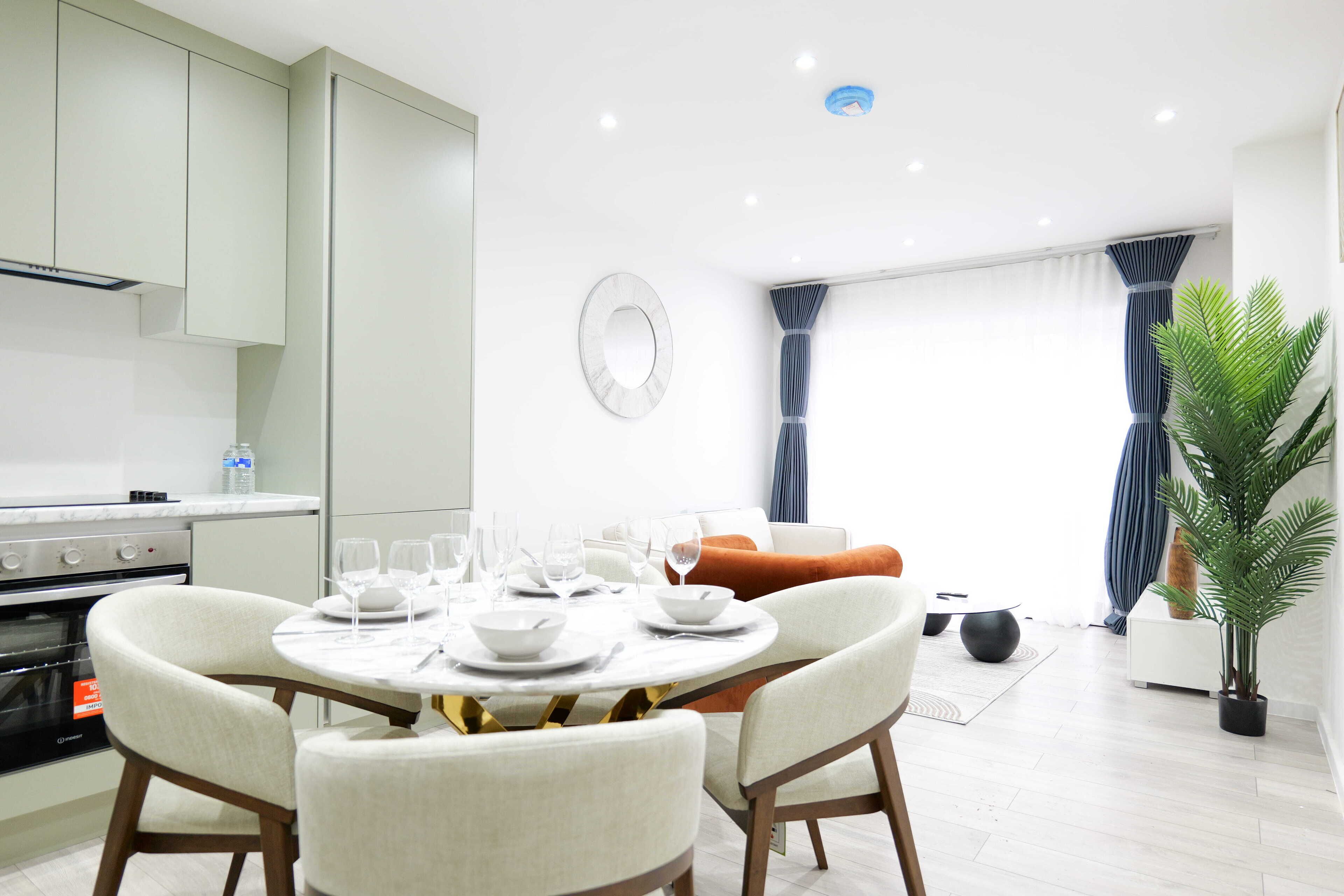 Une cuisine moderne avec des meubles blancs, un coin repas comprenant une table ronde et quatre chaises, et un salon avec un canapé et des rideaux.