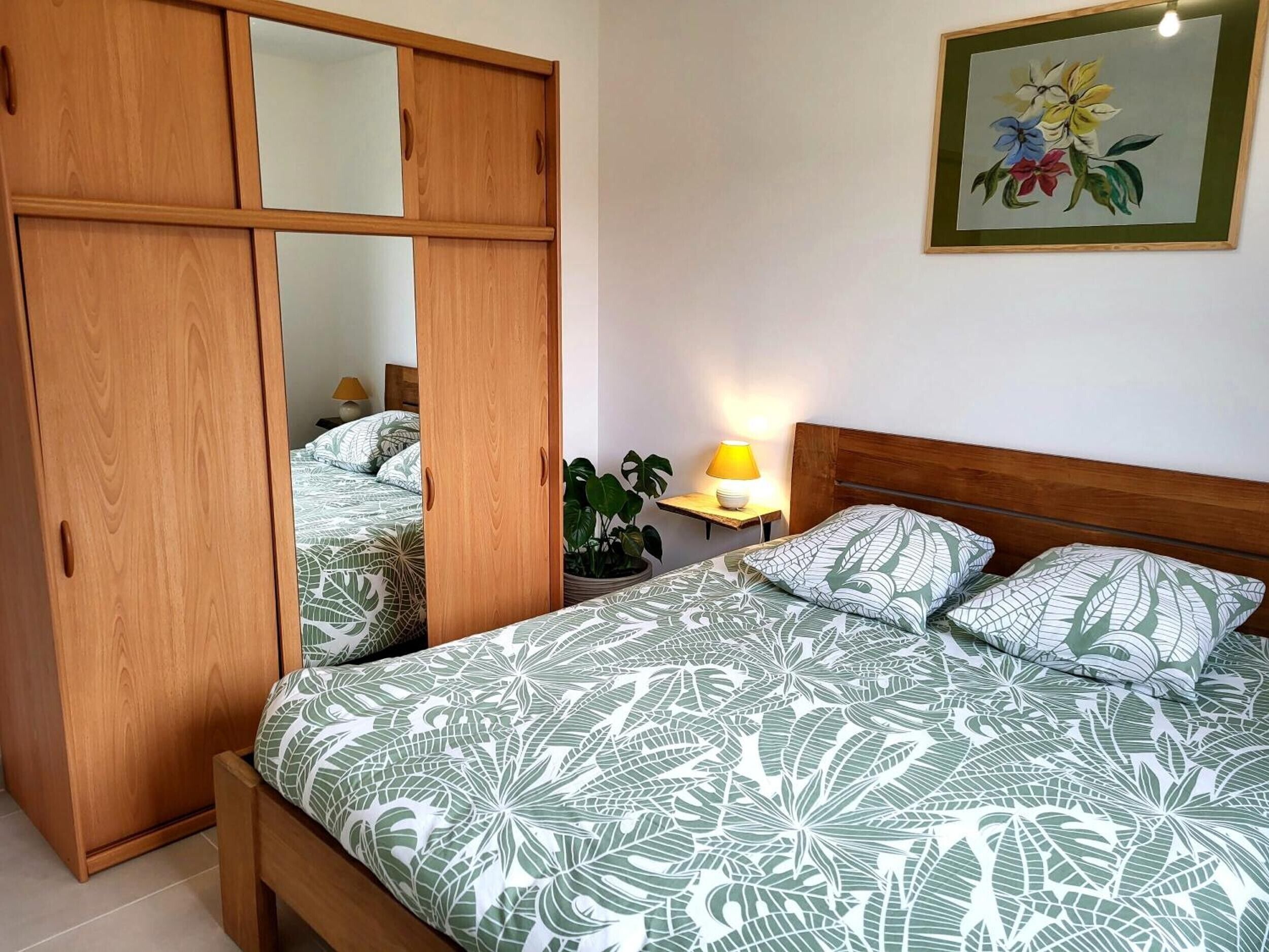 A bedroom with a bed, a wooden wardrobe, a nightstand with a lamp, and a wall-mounted painting.