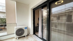 Un balcón con un equipo de aire acondicionado grande, una puerta corredera de vidrio y vista a un edificio de enfrente.