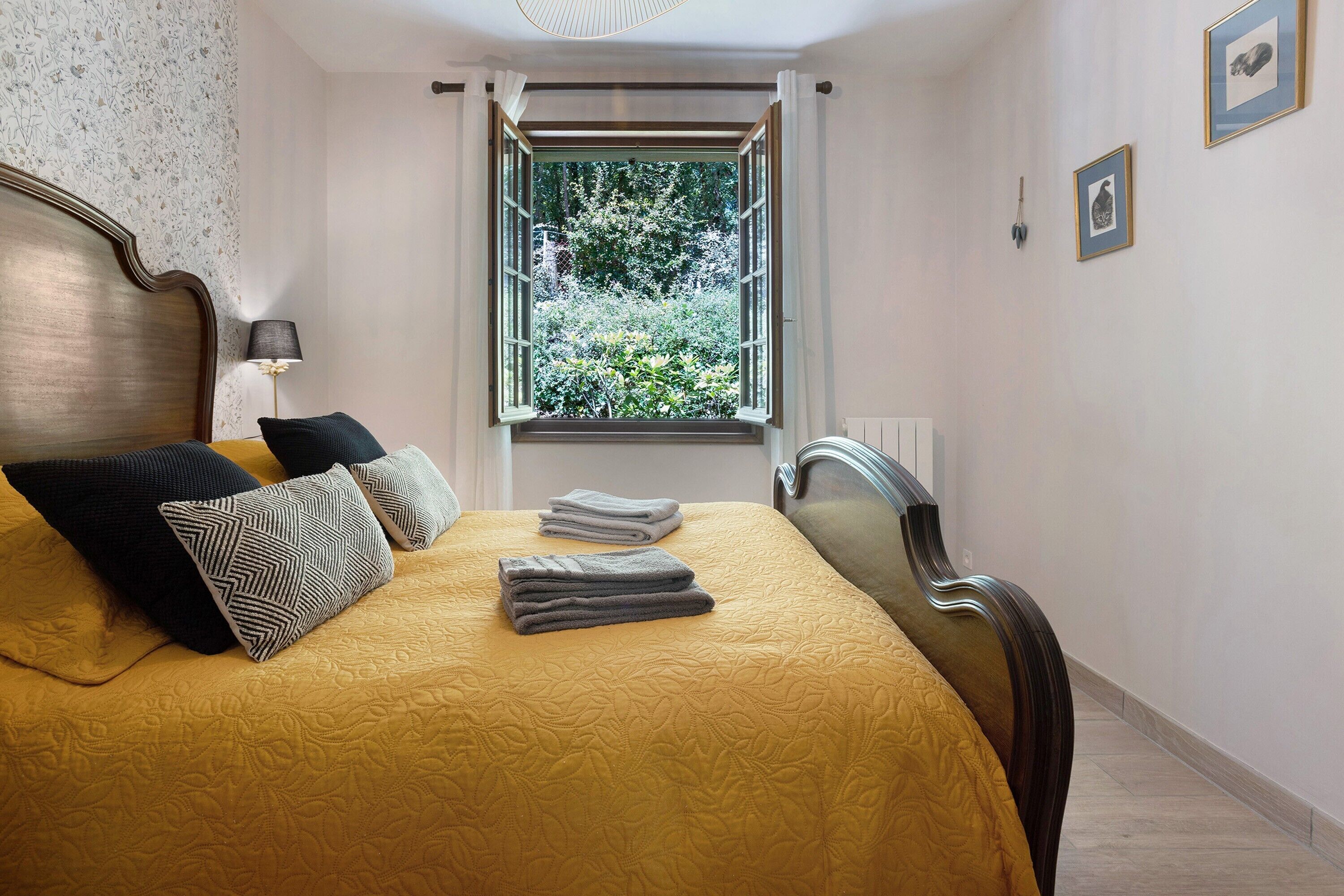 A neatly made bed with a yellow bedspread, pillows, and folded towels. A window with curtains is visible, and a lamp is on a bedside table.