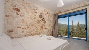 A bedroom with a stone wall, a bed with white linens, a view through a blue-framed window, and a hanging light fixture.