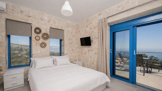Ein Schlafzimmer mit Bett, Fernseher, Fenster mit Aussicht und Balkon mit Stühlen.