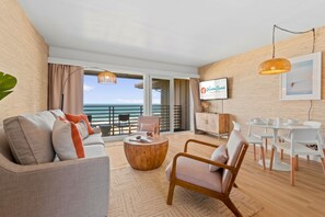 A modern living room with a sofa, coffee table, and dining area, featuring a large window with a view of the sea. - The Beach Bungalow | Ocean Front (San Diego)
